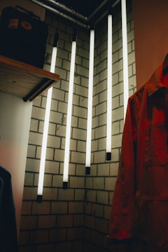 Vertical fluorescent light tubes hang from the ceiling against a backdrop of a white tiled wall. A wooden shelf is on the left, holding a black backpack. On the right, part of a coat or jacket is visible. The setting has an industrial or urban vibe.