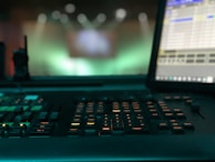 A dimly lit control panel with numerous buttons and keys is prominently featured, with a computer monitor displaying a program interface on the right. The background is blurred, with green and blue light bokeh, suggesting a focus on the equipment in a technological or production environment.