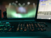 A dimly lit control panel with numerous buttons and keys is prominently featured, with a computer monitor displaying a program interface on the right. The background is blurred, with green and blue light bokeh, suggesting a focus on the equipment in a technological or production environment.