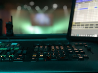 A dimly lit control panel with numerous buttons and keys is prominently featured, with a computer monitor displaying a program interface on the right. The background is blurred, with green and blue light bokeh, suggesting a focus on the equipment in a technological or production environment.