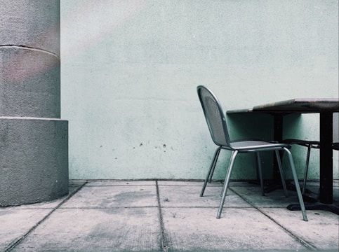 A minimalist scene featuring a metal chair placed beside a table against a plain light green wall. The setting is outdoors, possibly a patio, as indicated by the concrete paving. A large cylindrical pillar is seen to the left, adding a structural element to the composition.