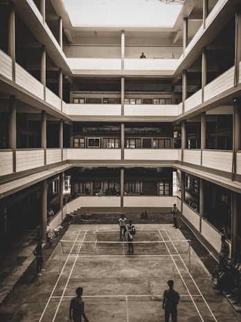 A multi-story building with an open courtyard features a concrete badminton court at the center. Several people are engaged in a game of badminton while others are watching from the sidelines. The architecture is utilitarian with balconies on each floor overlooking the courtyard.