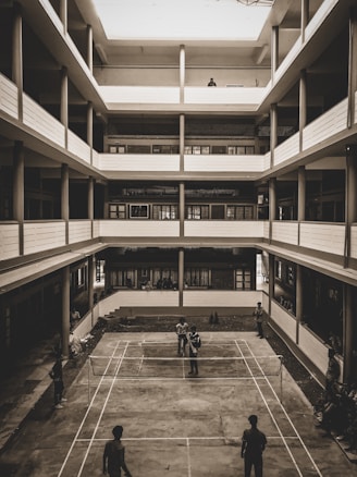 A multi-story building with an open courtyard features a concrete badminton court at the center. Several people are engaged in a game of badminton while others are watching from the sidelines. The architecture is utilitarian with balconies on each floor overlooking the courtyard.