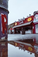 A close-up view of a painted metal surface, possibly part of a sculpture or structure, with a blurry background showing a street scene. The focus is on the peeling paint, with a reflection of buildings in a puddle on the ground. The street features colorful storefronts with signs, creating an urban atmosphere.