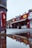 A close-up view of a painted metal surface, possibly part of a sculpture or structure, with a blurry background showing a street scene. The focus is on the peeling paint, with a reflection of buildings in a puddle on the ground. The street features colorful storefronts with signs, creating an urban atmosphere.