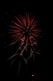 A single firework burst illuminates the night sky with vibrant red and white streamers radiating outward. The main burst features red spokes surrounded by a halo of sparkling white trails, creating a dramatic visual effect against the dark background.