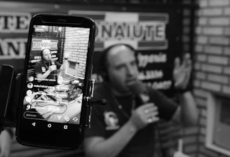 A podcast or radio host is seen speaking into a microphone, captured both directly and through the screen of a smartphone. The screen displays a live broadcast view with social media interactions. The background includes blurred signage, and the setting appears to be a studio with brick walls.