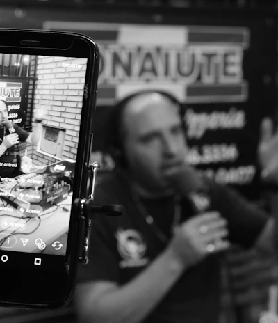 A podcast or radio host is seen speaking into a microphone, captured both directly and through the screen of a smartphone. The screen displays a live broadcast view with social media interactions. The background includes blurred signage, and the setting appears to be a studio with brick walls.