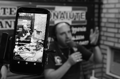 A podcast or radio host is seen speaking into a microphone, captured both directly and through the screen of a smartphone. The screen displays a live broadcast view with social media interactions. The background includes blurred signage, and the setting appears to be a studio with brick walls.