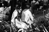 A black and white photograph featuring several people sitting on bicycles in an urban setting. The focus is on two women with braided hair, one wearing a floral dress and the other a striped tank top. Bicycles with baskets are positioned in the foreground, while more people walk in the background, creating a busy street atmosphere.