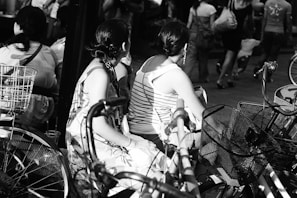 A black and white photograph featuring several people sitting on bicycles in an urban setting. The focus is on two women with braided hair, one wearing a floral dress and the other a striped tank top. Bicycles with baskets are positioned in the foreground, while more people walk in the background, creating a busy street atmosphere.