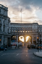A historical archway with an inscription in Latin at the top is framed against a sunset sky. The archway stands amidst classic, ornate buildings, and several vehicles and pedestrians are present on the street below. The street is dimly lit by streetlights, and the soft, warm glow of the setting sun can be seen through the arches.