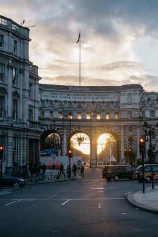 A historical archway with an inscription in Latin at the top is framed against a sunset sky. The archway stands amidst classic, ornate buildings, and several vehicles and pedestrians are present on the street below. The street is dimly lit by streetlights, and the soft, warm glow of the setting sun can be seen through the arches.