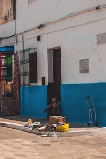 A street scene featuring a building with peeling paint and a bright blue lower facade. Various items, including textiles and packaged goods, are displayed on the ground along a sidewalk. A person is sitting in a doorway, partially obscured, adding to the everyday street life ambiance. A wheeled cart stands nearby, indicating a market or vendor setup.