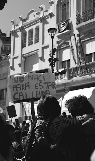 A black and white image of a city street scene with a crowd of people. They are holding signs, one of which reads 'No nací para estar callada.' The backdrop includes a large historic building with ornate architectural details. The scene is lively and appears to be part of a protest or demonstration.