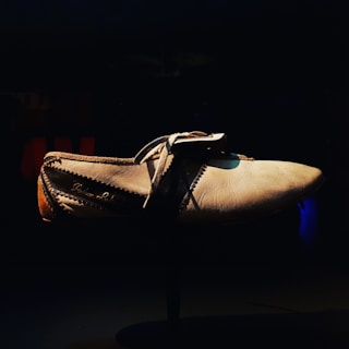 A single, worn shoe is positioned centrally in the frame, illuminated against a dark background. The shoe appears aged, with visible stitching and what looks like a lace tied over the top. The texture of the leather is highlighted by the lighting, giving it an old, used appearance.
