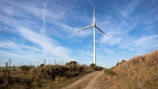 A tall wind turbine stands in a grassy open area under a bright blue sky with scattered clouds. The landscape includes a dirt path leading up to the turbine, surrounded by grassy hills and a few bushes. There is a fence running alongside the path.