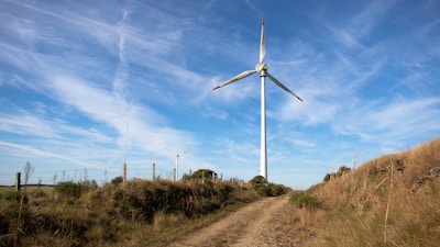 A tall wind turbine stands in a grassy open area under a bright blue sky with scattered clouds. The landscape includes a dirt path leading up to the turbine, surrounded by grassy hills and a few bushes. There is a fence running alongside the path.