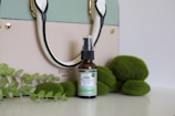 A stylish handbag in pastel colors is placed on a white background, complemented by a bottle of hemp oil positioned in front. Surrounding the oil bottle are decorative green elements resembling leaves and round moss-like objects, adding a natural touch to the composition.