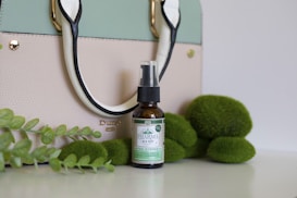 A stylish handbag in pastel colors is placed on a white background, complemented by a bottle of hemp oil positioned in front. Surrounding the oil bottle are decorative green elements resembling leaves and round moss-like objects, adding a natural touch to the composition.