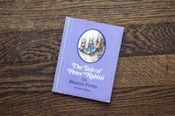 A book titled 'The Tale of Peter Rabbit' by Beatrix Potter is placed on a wooden surface. The cover is a light purple color featuring an illustration of a rabbit in a blue jacket. The text on the cover is positioned below the illustration.