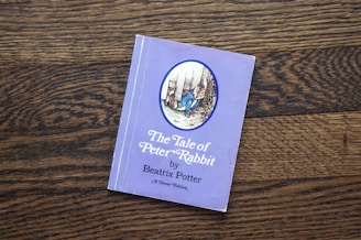 A book titled 'The Tale of Peter Rabbit' by Beatrix Potter is placed on a wooden surface. The cover is a light purple color featuring an illustration of a rabbit in a blue jacket. The text on the cover is positioned below the illustration.