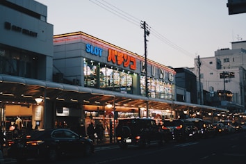 A bustling urban street scene with a brightly lit building featuring numerous signs, including one with 'SLOT' in large letters. The street is lined with cars and pedestrians, and the sky is a pale blue, indicating either early morning or early evening.