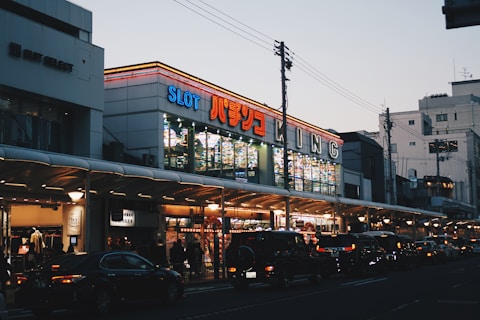 A bustling urban street scene with a brightly lit building featuring numerous signs, including one with 'SLOT' in large letters. The street is lined with cars and pedestrians, and the sky is a pale blue, indicating either early morning or early evening.