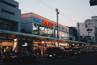 A bustling urban street scene with a brightly lit building featuring numerous signs, including one with 'SLOT' in large letters. The street is lined with cars and pedestrians, and the sky is a pale blue, indicating either early morning or early evening.