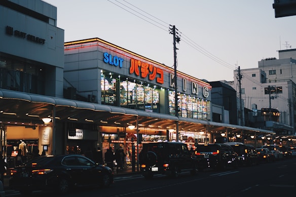 A bustling urban street scene with a brightly lit building featuring numerous signs, including one with 'SLOT' in large letters. The street is lined with cars and pedestrians, and the sky is a pale blue, indicating either early morning or early evening.