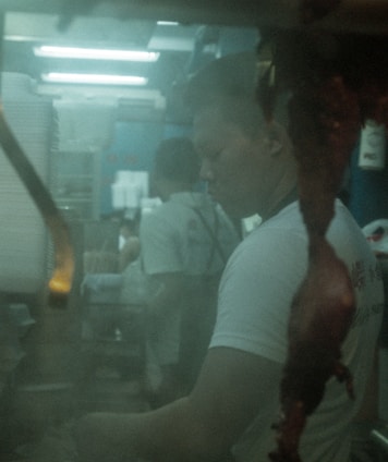 A person appears to be working in a kitchen environment with dim, soft lighting creating an atmospheric effect. The foreground features what seems to be a hanging piece of meat. The scene is partially blurred, contributing to a mysterious and introspective mood.