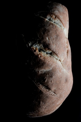 A loaf of rustic bread with a dark, crusty exterior is highlighted against a black background. The bread has a rough texture with several slashes across the top, suggesting it was baked with care. The lighting accentuates its artisanal qualities.