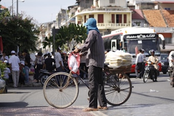 A person stands beside a bicycle on a bustling street. The bicycle has bags stacked on the rear. Nearby, a large coach bus and motorbikes are visible with people milling about, suggesting a lively urban environment. Buildings with varied architecture are in the background.