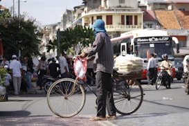 A person stands beside a bicycle on a bustling street. The bicycle has bags stacked on the rear. Nearby, a large coach bus and motorbikes are visible with people milling about, suggesting a lively urban environment. Buildings with varied architecture are in the background.