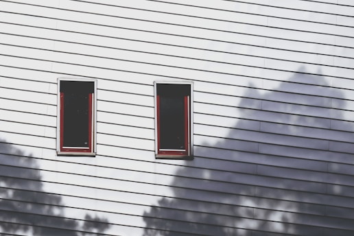 A wall with white horizontal siding features two rectangular windows with red trim. Shadows of trees are cast onto the wall, creating an interesting play of light and shape.
