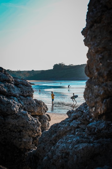 A coastal scene framed by rocky formations with people enjoying activities on a sandy beach. A person is holding a surfboard while others wade in the shallow waters. The sky is clear, and the distant landscape features some foliage.