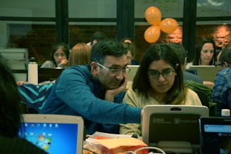 A group of people are working together in a collaborative environment. They are focused on their laptops, possibly engaged in a workshop or coding session. The room is softly lit with warm tones, and a few orange balloons are visible in the background.