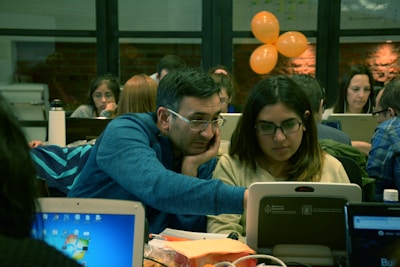 A group of people are working together in a collaborative environment. They are focused on their laptops, possibly engaged in a workshop or coding session. The room is softly lit with warm tones, and a few orange balloons are visible in the background.