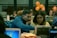 A group of people are working together in a collaborative environment. They are focused on their laptops, possibly engaged in a workshop or coding session. The room is softly lit with warm tones, and a few orange balloons are visible in the background.