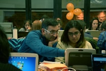 A group of people are working together in a collaborative environment. They are focused on their laptops, possibly engaged in a workshop or coding session. The room is softly lit with warm tones, and a few orange balloons are visible in the background.