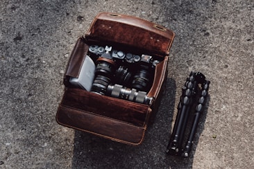 A vintage brown leather camera bag is open on a textured concrete surface, revealing a retro-style camera and several camera lenses inside. A compact black tripod is placed neatly beside the bag.