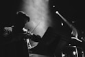 A silhouetted figure playing a violin under dramatic stage lighting, with visible smoke or fog adding to the atmosphere. The musician is intently focused on the music stand in front of them.