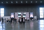 A large indoor event space with high ceilings, where a group of people are gathered around a stage area. The stage has a large screen and a podium, and several people are seated facing it. Others are standing or walking around the area. Tall windows line the walls, and large posters or banners are displayed on either side of the stage.