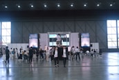 A large indoor event space with high ceilings, where a group of people are gathered around a stage area. The stage has a large screen and a podium, and several people are seated facing it. Others are standing or walking around the area. Tall windows line the walls, and large posters or banners are displayed on either side of the stage.