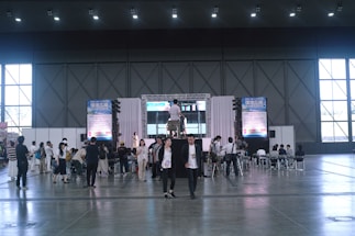 A large indoor event space with high ceilings, where a group of people are gathered around a stage area. The stage has a large screen and a podium, and several people are seated facing it. Others are standing or walking around the area. Tall windows line the walls, and large posters or banners are displayed on either side of the stage.