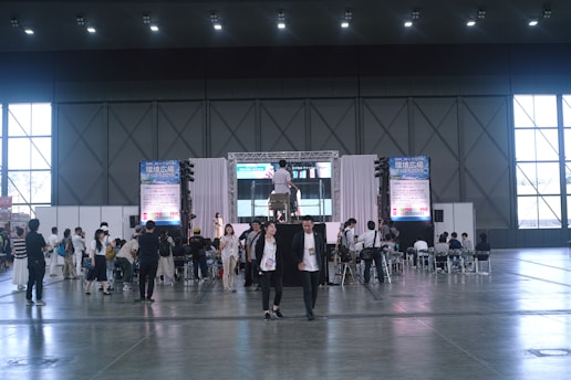 A large indoor event space with high ceilings, where a group of people are gathered around a stage area. The stage has a large screen and a podium, and several people are seated facing it. Others are standing or walking around the area. Tall windows line the walls, and large posters or banners are displayed on either side of the stage.