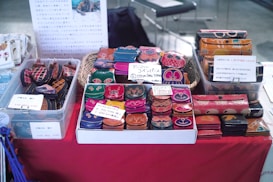 The table is covered with a variety of colorful, hand-crafted coin purses and eyeglass cases displayed in baskets and trays. Each item features vibrant designs with animal faces, patterns, and motifs. Some signage is visible with text in Japanese, providing prices and product details.