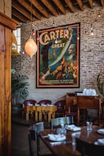 A vintage-style poster featuring a magician named Carter is prominently displayed on a rustic brick wall. The poster includes vibrant colors and dramatic illustrations of magical scenes. Below the poster, there's a cozy seating area with wooden tables and red chairs. Overhead, exposed wooden beams and string lights add to the ambiance. A stack of white napkins is neatly arranged on a wooden cabinet beside the seating area. A leafy plant is placed in the corner, adding a touch of greenery.