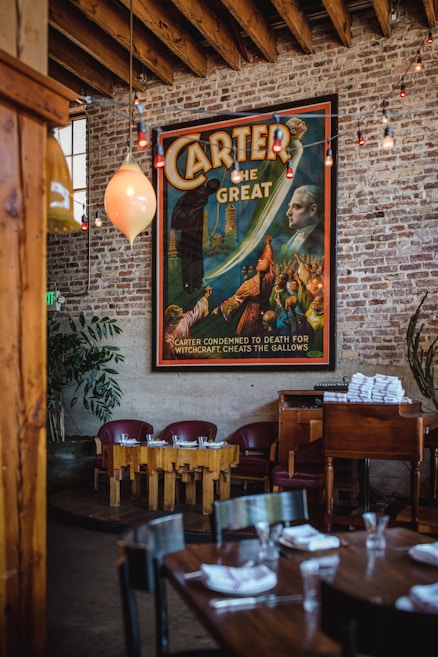 A vintage-style poster featuring a magician named Carter is prominently displayed on a rustic brick wall. The poster includes vibrant colors and dramatic illustrations of magical scenes. Below the poster, there's a cozy seating area with wooden tables and red chairs. Overhead, exposed wooden beams and string lights add to the ambiance. A stack of white napkins is neatly arranged on a wooden cabinet beside the seating area. A leafy plant is placed in the corner, adding a touch of greenery.