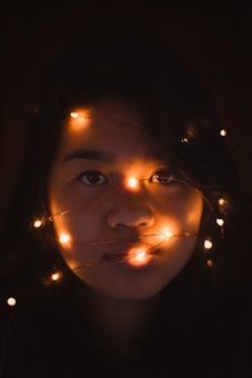 A person's face is softly illuminated by strands of warm, glowing fairy lights. The surrounding area is dark, creating a contrast with the gentle light on the skin.